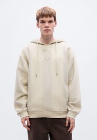 Φούτερ με κουκούλα σε εκρού χρώμα με κάθετη ριμπ υφή, κουκούλα με κορδόνι και μικρό λογότυπο σε σχήμα ασπίδας στο στήθος. Άνετη εφαρμογή με μακριά μανίκια.