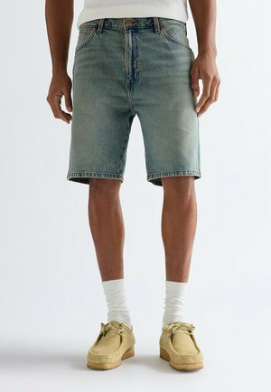 Uomo che indossa pantaloncini di jeans blu sbiaditi, calzini bianchi, mocassini color cuoio e una camicia bianca, in piedi contro uno sfondo semplice.