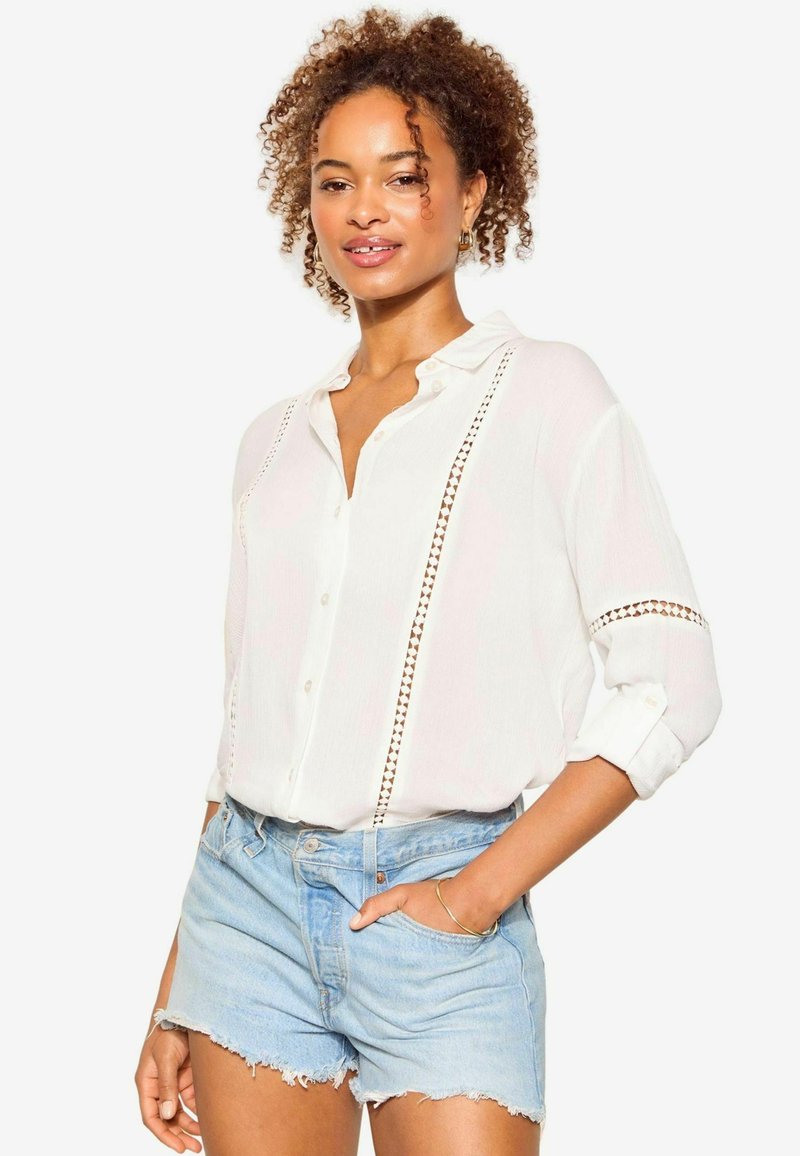 Camicia bianca con accenti in pizzo, maniche lunghe arrotolate fino ai gomiti, abbinata a pantaloni shorts in denim azzurro chiaro. Tessuto liscio e vestibilità rilassata.