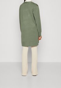 Kvinna iklädd en lång olivgrön cardigan över en vit skjorta med krage och krämfärgade ribbade vida byxor, stående mot en neutral bakgrund.