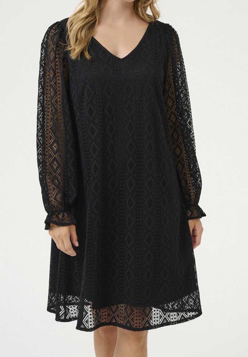 Femme portant une robe en dentelle noire à motifs géométriques, longueur genou, avec des manches longues transparentes, debout devant un fond clair et uni.