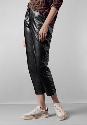 Schwarze Kunstlederhosen mit lockerer Passform, ausgestattet mit einem elastischen Bund und sichtbaren Nähdetails, kombiniert mit beigen Sneakers.