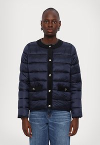 Navy puffer jacket with horizontal quilted design, black accents, snap buttons, and two front pockets. Worn over a gray sweater.