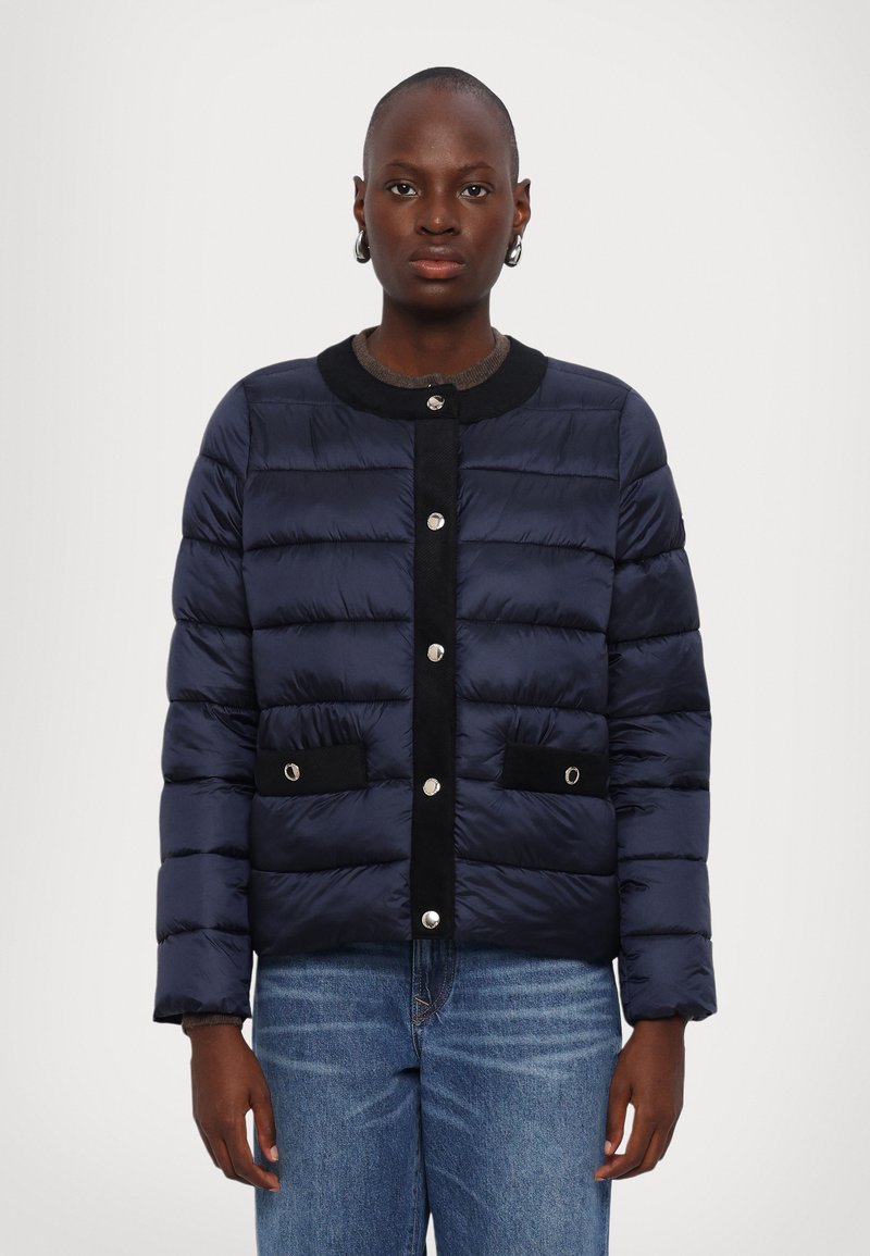 Navy puffer jacket with horizontal quilted design, black accents, snap buttons, and two front pockets. Worn over a gray sweater.