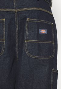 Jeansoveralls i mørk indigo med gule syninger. Har flere lommer og et Dickies-logo på den højre lomme.