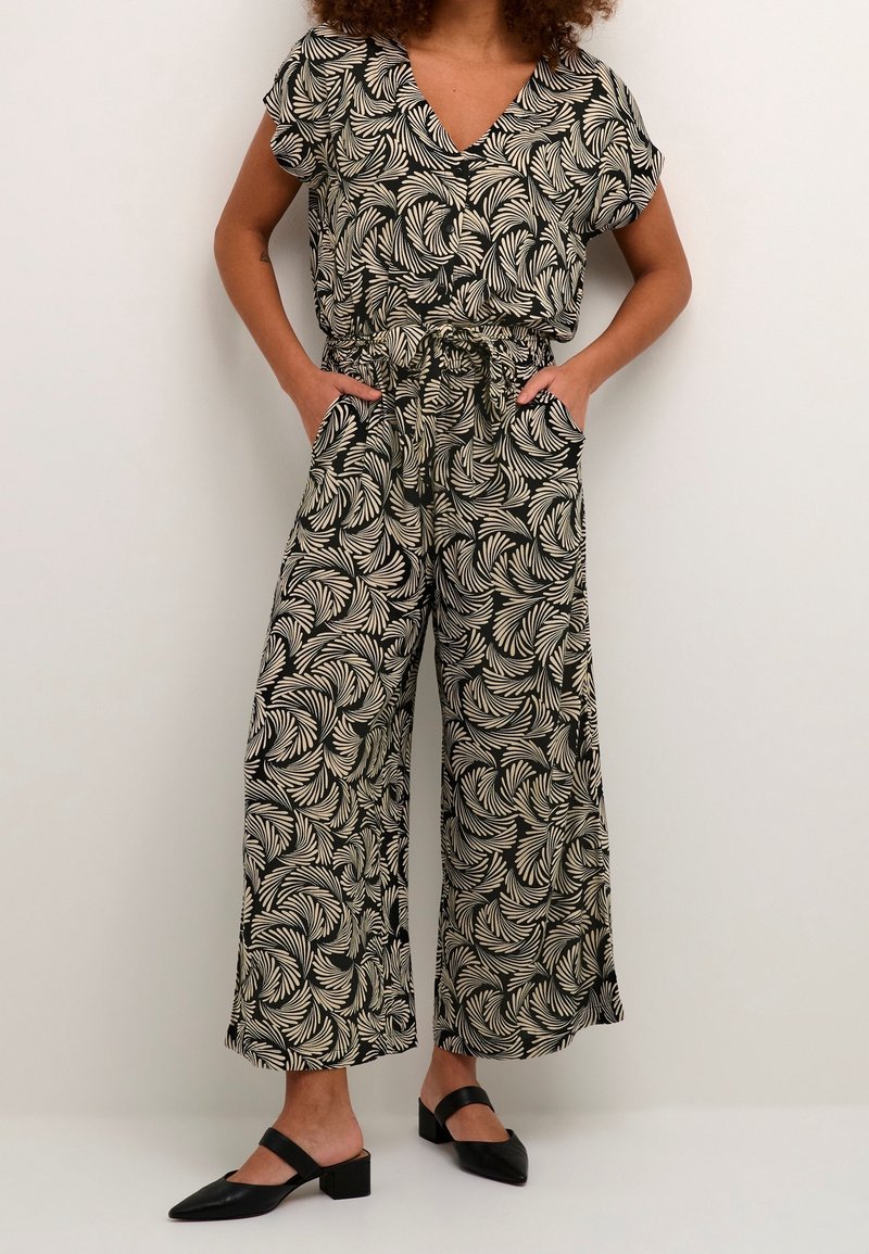 Femme portant une combinaison à manches courtes à col en V avec motif noir et beige, pantalon à jambes larges et chaussures noires à petit talon, mains dans les poches.