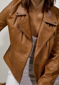 Giacca da moto in pelle marrone con accenti trapuntati, zip argentate e bottoni a pressione. Vestibilità su misura con ampie rever e finitura testurizzata.