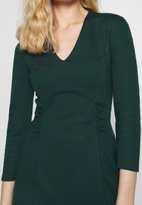 Green dress made of smooth fabric, featuring a V-neck, three-quarter sleeves, and gathered detailing on sides for a fitted silhouette.