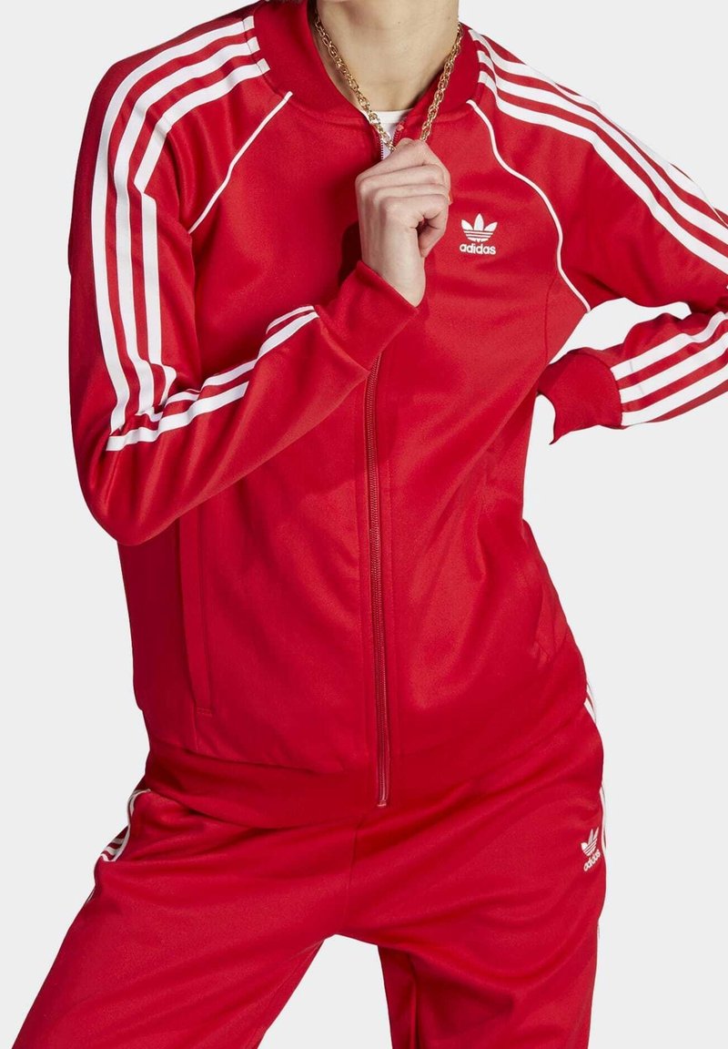 Röd polyester-tracksuit med vita ränder längs axlarna och sidorna. Har dragkedja framtill och logotyp på bröstet. Slät textur.