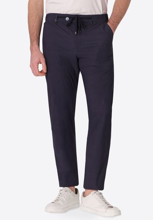 Uomo che indossa pantaloni slim fit blu navy con vita a coulisse e sneakers bianche, in piedi con una mano in tasca e una mano rilassata.
