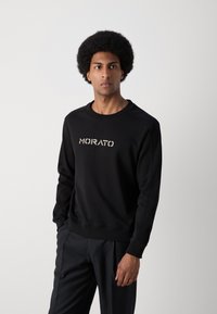 Sudadera negra con puños y dobladillo acanalados, que presenta el logo "MORATO" en un color contrastante. Llevada con pantalones oscuros de corte entallado.