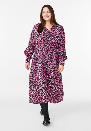 Långärmad, knälång klänning i rosa och svart leopardmönster med bälte i midjan, flödande tyg och V-ringning. Bärs över svarta strumpbyxor.