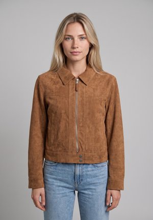 Donna bionda che indossa una giacca scamosciata marrone con zip e collo, abbinata a jeans azzurri, in piedi davanti a uno sfondo grigio uniforme.