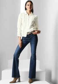 White button-up shirt with lace trim, paired with dark blue flared jeans and beige high-heeled shoes. Model stands on white steps.