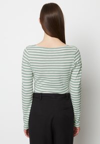 Frau mit langen, glatten braunen Haaren, die ein hellgrünes und weiß gestreiftes Langarmoberteil und schwarze hoch taillierte Hosen trägt und wegschaut.