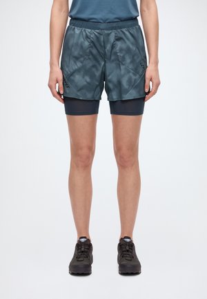 Un mannequin porte un short de running sombre à motifs par-dessus un short de compression ajusté, avec des chaussures de running noires, sur un fond blanc uni.