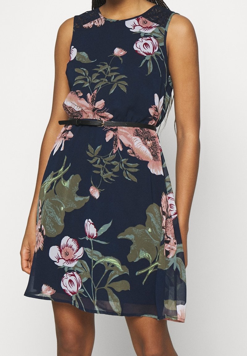 Robe florale en tissu bleu marine. Présente des motifs floraux roses et verts, un design sans manches et une ceinture noire à la taille.