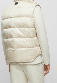 Light beige padded vest with a subtle diamond pattern, featuring a high collar and a logo tag on the back, paired with a light sweater.