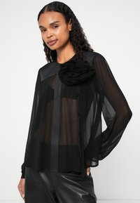 Blusa nera trasparente con maniche lunghe, caratterizzata da un dettaglio floreale arricciato sulla spalla e righe verticali. Tessuto morbido e vestibilità rilassata.