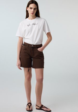 Mujer con camiseta blanca "Warm Heart", pantalones cortos marrones con cinturón y sandalias marrón oscuro, de pie con una mano en el bolsillo frente a un fondo liso.