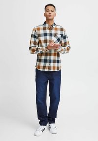 Camicia a maniche lunghe a quadri in marrone, bianco e blu navy, con taschino sul petto, abbinata a jeans blu scuro e sneaker bianche.