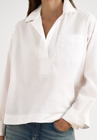 Valge puuvillane pluus, millel on kraega kaelus, poolkukuga plastron ja rinnatasku. Pika varrukaga, varrukate all äärtega, lõdva lõikega.