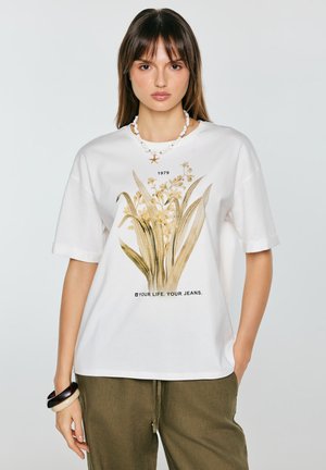 Naine kannab valget t-särki beeži lillemustri ja tekstiga, oliivrohelisi pükse, kestkaelakeed tähtkalakujulise ripatsiga ning käevõrusid vasakul randmel.