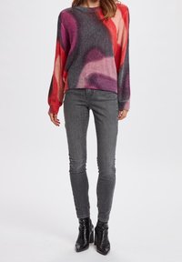 Pull en tricot multicolore avec des nuances de rose, gris et rouge ; coupe décontractée ; associé à un jean gris ajusté et des bottes noires à talons hauts.