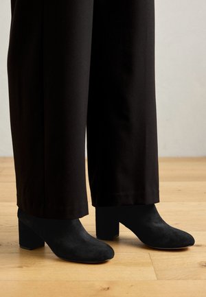 Schwarze Wildleder-Stiefeletten mit abgerundeter Spitze und einem blockförmigen Absatz, kombiniert mit schwarzen weiten Hosen auf einem Holzboden.