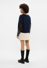 Granatowy sweter wełniany o teksturowanym wykończeniu, z wielokolorowymi rękawami w patchworkowym stylu oraz luźnym kroju, zestawiony z kremową spódnicą i czarnymi botkami.