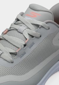 Lys grå atletisk sko med teksturert mesh-overdel, tykke lisser og korallfarget logo på polstret tunge.