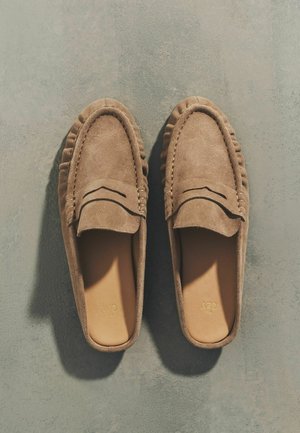 Para jasnobrązowych mokasynów zamszowych na wsuwkę z detalami przeszyć, ułożonych obok siebie na szarej, teksturowanej powierzchni.