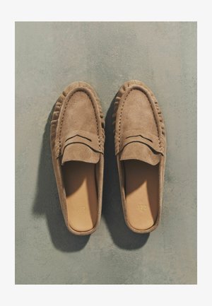 Para jasnobrązowych mokasynów zamszowych na wsuwkę z detalami przeszyć, ułożonych obok siebie na szarej, teksturowanej powierzchni.