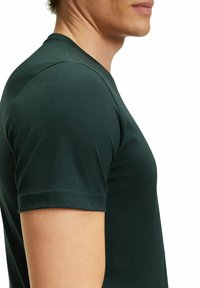 T-shirt verde scuro a maniche corte realizzato in morbido cotone, con un design aderente e dettagli di cucitura sottili lungo i bordi.