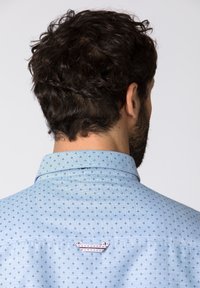 Muž s kadeřavými tmavými vlasy, oblečený v světle modré vzorované košili, zobrazený zezadu na jednobarevném světlém pozadí.