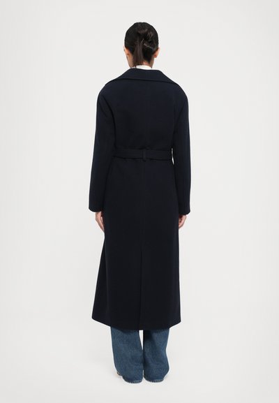 Donna in piedi con la schiena rivolta verso la macchina fotografica, che indossa un lungo cappotto blu navy con cintura e jeans blu su uno sfondo bianco semplice.
