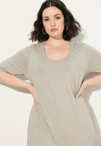 Camiseta de color beige claro con escote barco, hecha de una tela suave, con mangas cortas y un corte relajado. Destaca por sus bolsillos laterales y su diseño sencillo.