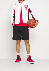 Hvid varsityjakke med røde og sorte accenter, rød t-shirt, sorte shorts og røde basketballsko; holder en basketball.