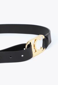 Svart läderbälte med en guldmetallspänne som har en utskuren logodesign; slät yta med flera justeringshål.