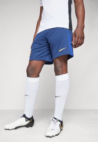 Nike futbols šorti zilā krāsā ar zeltītu logotipu, apvienoti ar baltām zeķēm un melnbaltiem apaviem. Izstrādāti sportiskai veiktspējai.