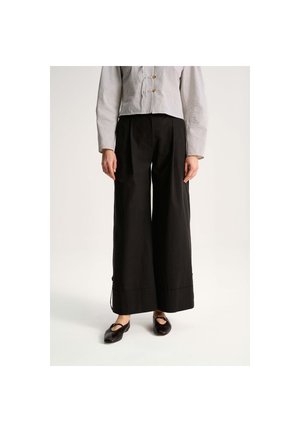 Osoba nosząca czarne spodnie z szerokimi nogawkami, krótki, kraciasty żakiet z guzikami na zatrzask oraz czarne baleriny, stojąca na jednolitym tle.