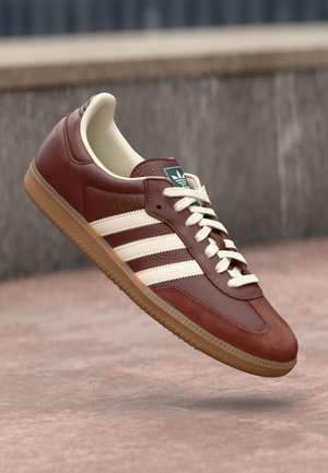Bruine Adidas Samba sneaker met witte strepen en veters, groen logo op de tong en een gum rubberen zool, zwevend in de lucht tegen een stedelijke achtergrond.
