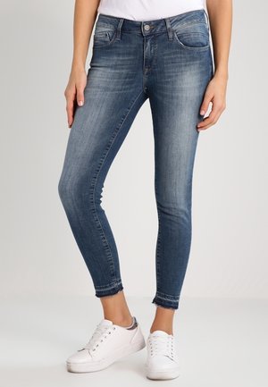 Slim fit blå jeans med falmet vask, slidte kanter, båret med hvide sneakers og en hvid top, vist på en person der står.