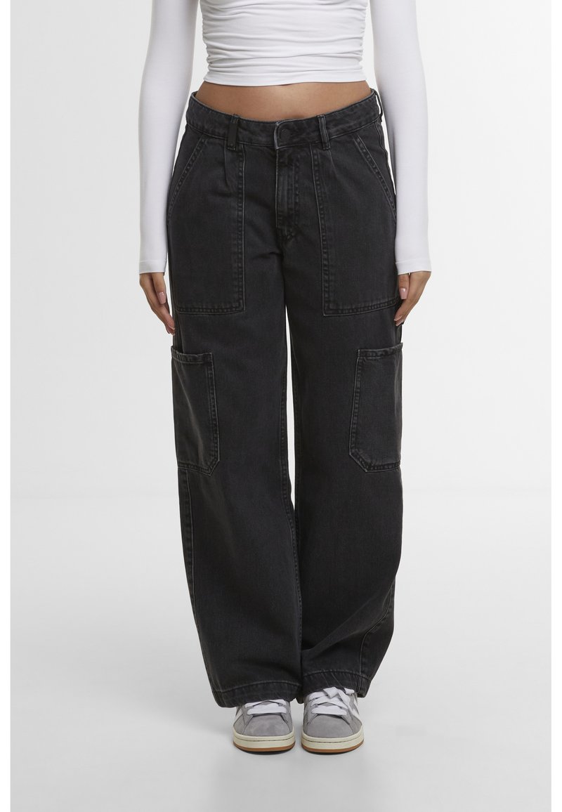 Femme portant un jean cargo noir à jambes larges avec de grandes poches avant et latérales, un crop top blanc à manches longues et des baskets grises.