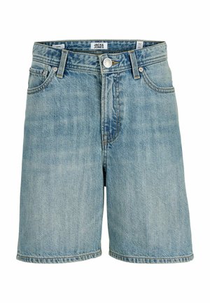 Poika, jolla on vaaleansininen lippalakki, vaaleanpunainen t-paita, farkkushortsit, valkoiset sukat ja lenkkarit, seisoo ja katsoo sivulle yksivärisellä taustalla.