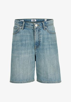 Dreng iført en lyseblå kasket, lyserød T-shirt, cowboyshorts, hvide sokker og sneakers, der står og ser til siden på en ensfarvet baggrund.