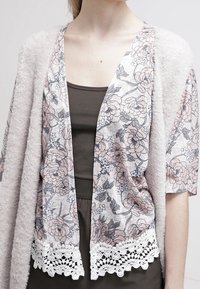 Offene Strickjacke mit weicher rosa Textur, floral gemustertem Innenstoff und weißen Spitzenbesatz am Saum. Kurze Ärmel.