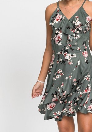 Femme portant une robe verte à fleurs sans manches avec des volants et des bretelles fines, montrée des épaules aux genoux sur fond blanc.