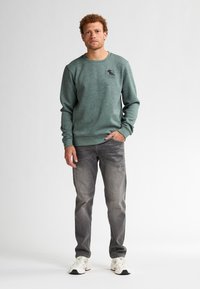Felpa verde con scollatura a girocollo e logo, abbinata a jeans grigi e sneaker bianche. Vestibilità casual e tessuto testurizzato.