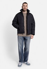 Black quilted puffer jacket with high collar and snap closure. Worn over a beige shirt, paired with blue jeans and sneakers.